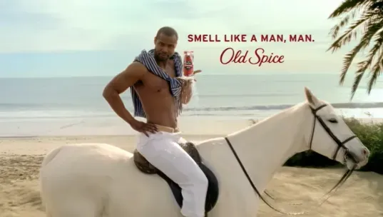 Исайя Мустафа в рекламе Old Spice сидит верхом на белой лошади с флаконом дезодоранта в руке